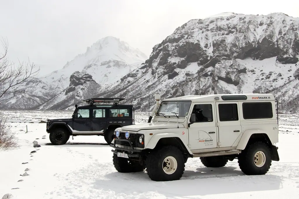 Two white MudShark Defenders parked on a snow-covered plain below a steep white mountain.