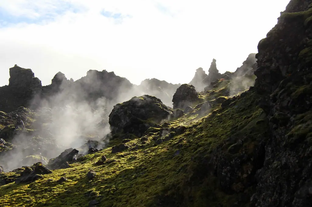 Geothermal steam rising between jagged lava spires above a slope of bright green moss.