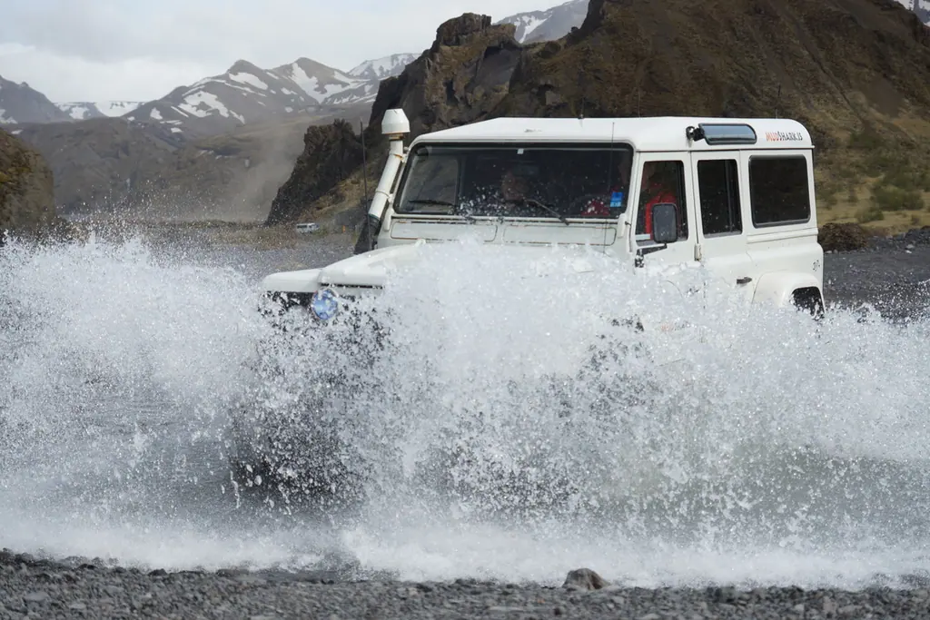 A white super-jeep driving through a glacial river, throwing up a wall of spray.
