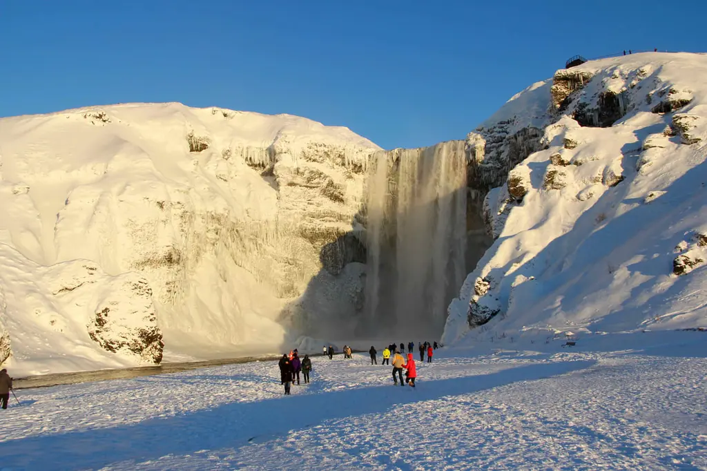 A wide waterfall falling between snow-covered slopes, with a line of people in bright jackets standing small at its foot.